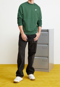 En person klädd i en grön Nike-tröja, svarta jeans och vita sneakers står bredvid ett grått tre-lådigt arkivskåp på ett gult golv.