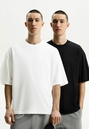 Due uomini, uno in una t-shirt di cotone bianca oversize e l'altro in un corrispondente nera, entrambi con capelli corti e sfondi neutrali.