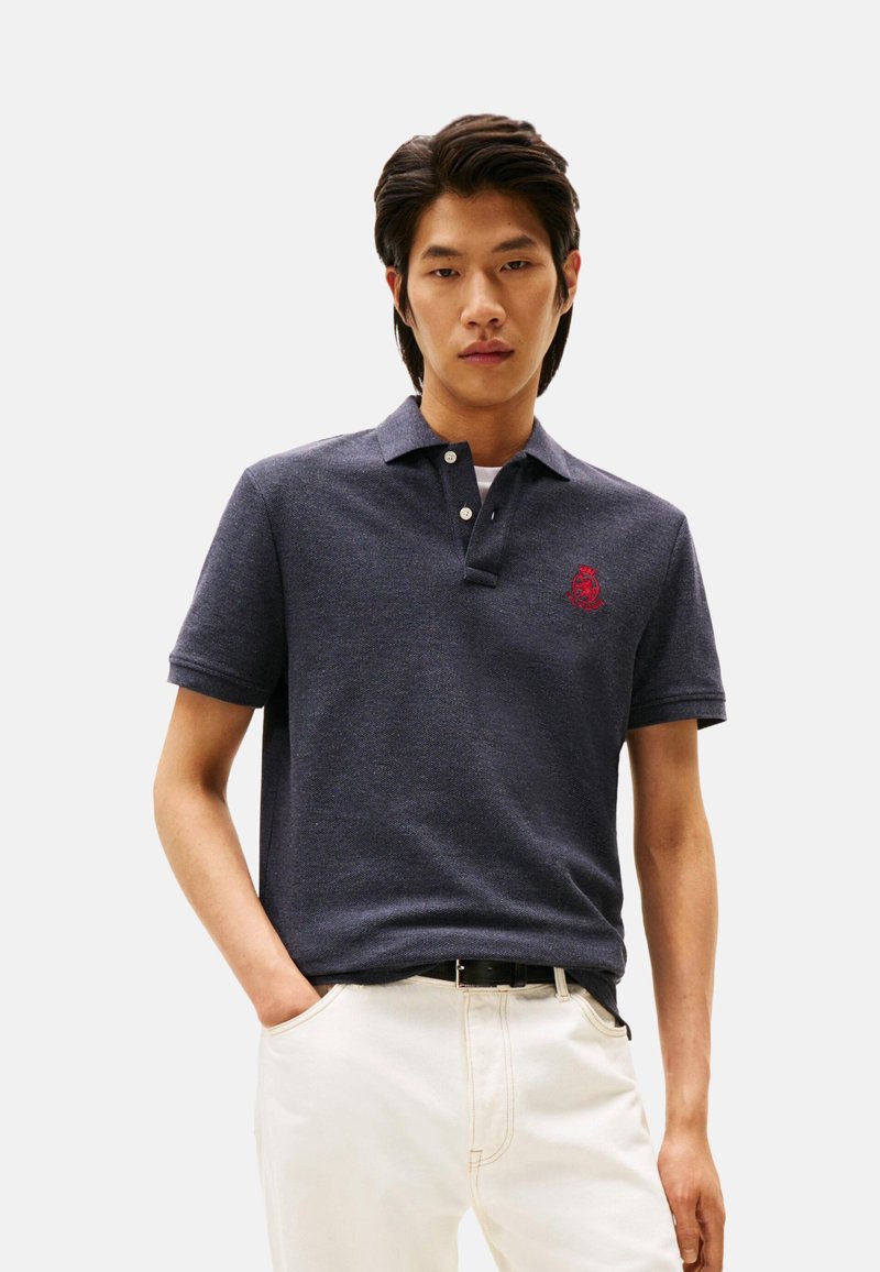 Polo de color azul marino con cuello abotonado, que presenta un logo rojo bordado en el pecho, combinado con pantalones de color claro. Textura suave.