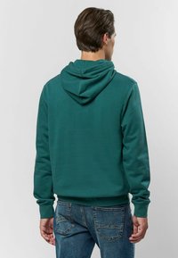 Zöld kapucnis pulóver, puha anyagból készült, tágas kapucnival, bordázott mandzsettákkal és derékrésszel. Farmernadrággal viselve, sima hátoldali dizájnnal.