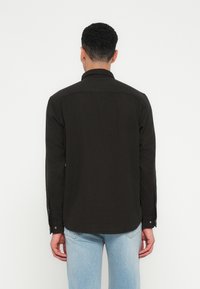 Hombre de espaldas vistiendo una camisa negra de manga larga y jeans de un azul claro, contra un fondo blanco liso.