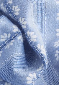 Tissu tissé bleu avec motifs floraux blancs et détails cousus, montré en gros plan avec des plis doux et une texture.