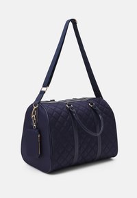 Marinblå quiltad duffelväska med dubbla handtag och en justerbar rem. Har en framficka med dragkedja och en accent av guldfärgad hårdvara.