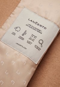 Etykieta z materiału wełnianego w delikatnym odcieniu brzoskwiniowym, z białym geometrycznym wzorem, zawierająca symbole pielęgnacji oraz logo "LANCERTO". Teksturowane tło.