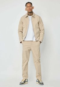 Jeune homme avec un tatouage au cou portant une veste beige, un pantalon beige, une chemise blanche et des baskets noir et blanc, se tenant devant un fond uni.