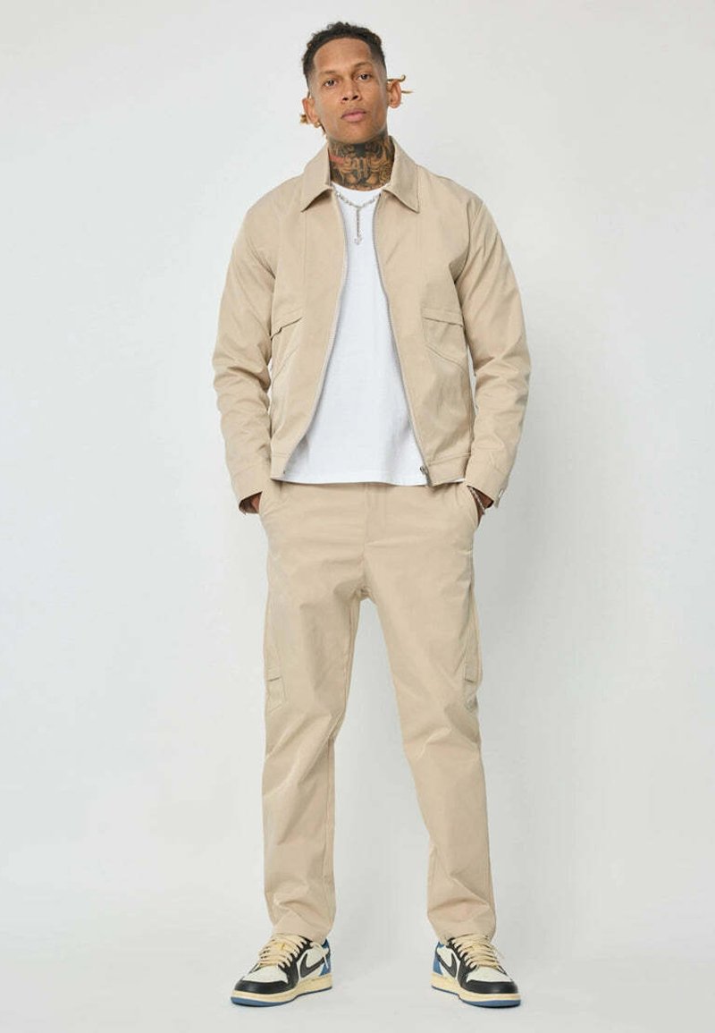 Jeune homme avec un tatouage au cou portant une veste beige, un pantalon beige, une chemise blanche et des baskets noir et blanc, se tenant devant un fond uni.