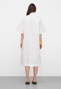 Woman standing with back visible, wearing a white loose knee-length dress with short sleeves, black heeled sandals, and a yellow bracelet.