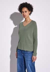Maglione verde a V con una leggera lucentezza, maniche lunghe e una vestibilità rilassata, abbinato a jeans a gamba larga blu. Tessuto morbido e design casual.