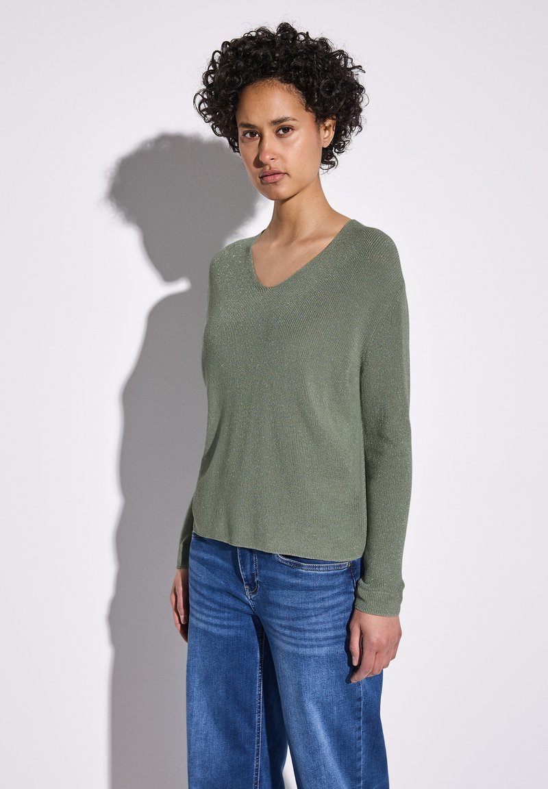 Maglione verde a V con una leggera lucentezza, maniche lunghe e una vestibilità rilassata, abbinato a jeans a gamba larga blu. Tessuto morbido e design casual.