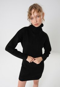 Crna mini haljina s visokim ovratnikom izrađena od mekog materijala. Ima dugačke rukave i rebraste manžete, dizajnirana je s uskim krojem za elegantnu siluetu.