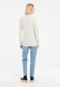 Pull en col roulé beige à côtes avec des manches longues, associé à un jean bleu clair et des bottines noires à semelles épaisses. Fond uni.