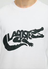 Chemise blanche avec un logo Lacoste noir combinant le nom de la marque et un graphisme stylisé d'alligator sur la poitrine.