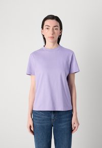 Vaalean violetin värinen puuvillapaita, jossa on pyöreä kaula-aukko, lyhyet hihat ja keskellä brodeerattu logo. Suora helma.