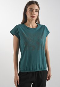 Modelka v tyrkysovém topu s kr�átkým rukávem a dekoračním vzorovaným designem. Pozadí je neutrální a nenápadné, zaměřuje se na outfit.