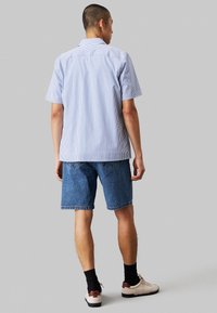 Camicetta a maniche corte, a righe blu e bianche abbinata a pantaloni shorts in denim. La camicia ha un colletto classico, mentre i pantaloni shorts sono al ginocchio e casual.