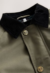 Veste vert olive avec un col en velours côtelé noir, un bouton en métal à motif flèche, et une texture de tissu lisse. Doublure intérieure à carreaux foncés visible.