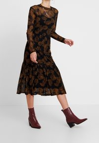 Svart klänning med långa genomskinliga ärmar, prydd med ett orange blommönster, en utsvängd fåll och bourgognefärgade klackstövlar som når ankeln.