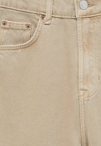 Beige farkut, joissa on klassinen viiden taskun muotoilu, kupariset niitit ja kontrastivärinen ompelu. Kankaan pinta on hienovaraisesti teksturoitu.