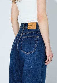 Jeans in denim scuro a vita alta con una patta in pelle sul retro, dotati di due tasche posteriori e cuciture a contrasto.