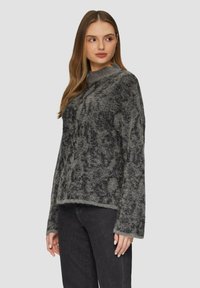 Pull gris flou avec un motif texturé et un léger brillantage. Il présente un col montant et des manches longues, associé à un jean foncé.
