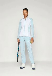 Lys blå idrettsjakke med hvite aksenter og glidelås, sammen med matchende bukser. Figuren holder en tennisracket og ball.