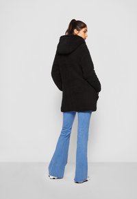 Veste à capuche noire en tissu pelucheux et texturé, dotée d'une coupe décontractée et de poches latérales, associée à un jean évasé bleu clair.