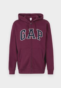 Sudadera con cremallera color granate hecha de tela suave, con un gran logo "GAP" en azul marino y blanco. Incluye capucha y bolsillos frontales.