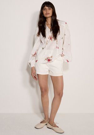 Femme aux longs cheveux foncés portant un chemisier blanc brodé de fleurs, un short blanc et des ballerines beige à lacets, debout devant un fond uni.