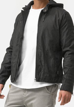 Mann trägt schwarze Lederjacke mit Reißverschluss und Kapuze, weißes T-Shirt und hellgraue Hose, steht vor einem einfarbigen Hintergrund.