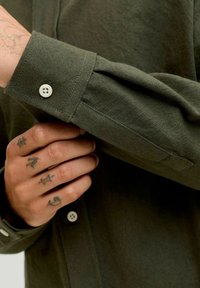 Oliivroheline nööpidega särk, millel on tekstureeritud kangas, üks valge nupp mansetil ning lähivõte käest, millel on tätoveeringud.