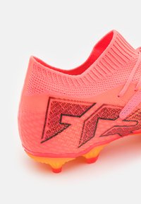 Felroze voetbalschoen met een gestructureerde breisel bovenkant, uitgevoerd met een opvallend zwart grafisch ontwerp en neongele buitenste studs voor grip.