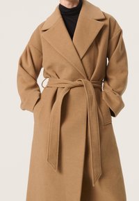 Wollmantel aus Kamelhaar mit Gürtel in der Taille, breiten Revers und langen Ärmeln. Verfügt über eine weiche Textur und seitliche Taschen.
