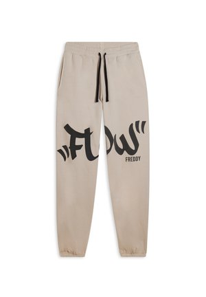 Pantalon de survêtement beige en tissu doux avec une taille à cordon de serrage, arborant un grand graphique noir "FLOW" et le texte "FREDDY" sur la jambe gauche.