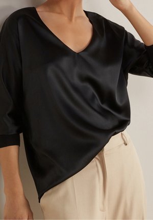 Kobieta w czarnej satynowej bluzce z dekoltem w serek i długimi rękawami, wpuszczonej w beżowe spodnie z wysokim stanem na neutralnym tle.