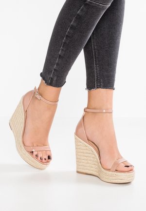 Woman wearing black jeans and beige ankle-strap wedge sandals with braided rope heel standing on a white surface.