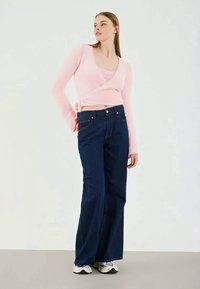 Suéter rosa felpudo em estilo envolvente, jeans de ganga de perna larga azul escuro, ténis brancos e cinzas. Texturas suaves e um ajuste descontraído, em pé sobre um fundo neutro.
