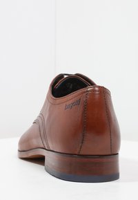 Chaussure habillée pour homme en cuir marron avec détails cousus, petit talon en bois, et logo « bugatti » sur le côté, vue de l'arrière.