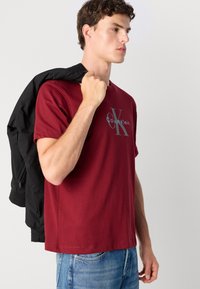 Bordo majica s kratkim rukavima s Calvin Klein logotipom; model nosi crnu bomber jaknu preko ramena. Klasičan kroj, pamučna tkanina.