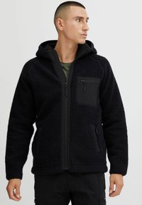 Chaqueta de forro polar negra con capucha, exterior texturizado, cierre frontal con cremallera y bolsillo en el pecho, y detalles en negro contrastante en los puños y bolsillos.