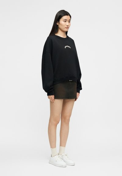 Γυναίκα που στέκεται φορώντας μαύρο φαρδύ φούτερ, σκούρα μίνι φούστα, λευκές κάλτσες και λευκά αθλητικά παπούτσια μπροστά σε απλό, ανοικτό φόντο.