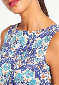 Beige mouwloze jurk met bloemmotief en blauwe en oranje accenten. Heeft een ronde halslijn en textiel met structuur.