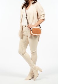 Borsa a tracolla in suede color tan con rifiniture in pelliccia sintetica color crema, forma rettangolare, chiusura con zip e tracolla regolabile. Modella in abbigliamento beige.