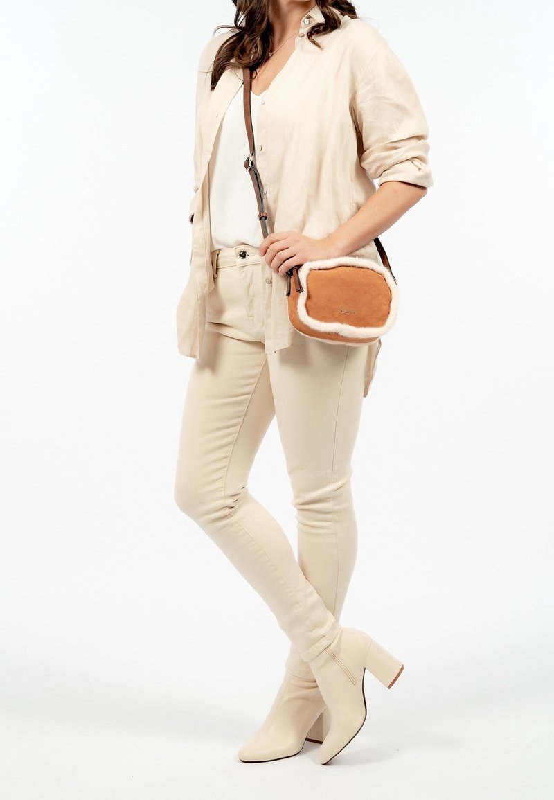 Borsa a tracolla in suede color tan con rifiniture in pelliccia sintetica color crema, forma rettangolare, chiusura con zip e tracolla regolabile. Modella in abbigliamento beige.