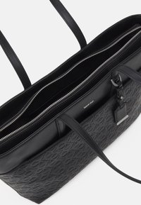 Černá kožená taška přes rameno s embosovaným vzorem, dvěma dlouhými uchy a zipy. Podšitý interiér s několika odděleními pro uspořádání.