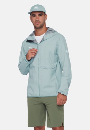 Uomo che indossa una giacca leggera azzurro chiaro con zip e un cappellino, insieme a pantaloncini verdi, mentre sistema la zip della giacca su uno sfondo neutro.