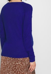 Blå stickad tröja med ribbade axlar, slim fit och långa ärmar, i kombination med en brun prickig kjol med vita prickar.