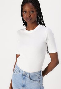 T-shirt bianco a righe con maniche corte e scollo rotondo, indossato con jeans a vita alta di colore blu chiaro, caratterizzato da un design minimali e una superficie liscia.