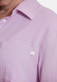 Lichtpaarse shirt gemaakt van gestructureerde stof, met een kraag, een borstzakje en een klein wit label aan de zijkant.