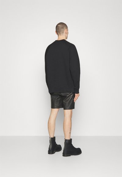 Sweatshirt noir à manches longues, associé à un short en cuir noir. Le mannequin porte des bottines noires à fermetures éclair sur le côté. Design épuré et minimaliste.