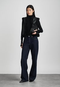 Zwarte leren vest met bontafwerking, coltrui, ruime donkere denim jeans en een kleine zwarte handtas. Model staat tegen een lichte achtergrond.